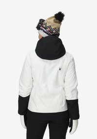 Veste de ski blanche et noire avec un col haut et un logo dans le dos, assortie d'un bonnet en tricot à motifs et de gants, présentant un design sportif.