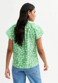 Blouse verte à fleurs avec des manches courtes bouffantes, présentant des fleurs blanches sur un fond lumineux. Le dos a un design froncé. Associée à un jean en denim clair.