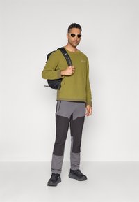 Pullover felpato verde, pantaloni tecnici grigi e neri con tasche zippate, zaino nero e occhiali da sole, tutti che mettono in mostra attrezzatura outdoor funzionale.