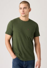 Tricou tricotat de culoare verde măslin, cu mâneci scurte, decolteu rotund și un buzunar pe piept. Prezintă o textură subtilă și eticheta logo-ului Levi's.