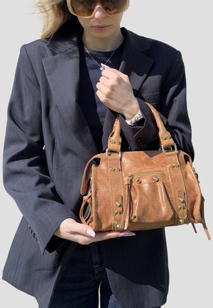 Donna con blazer scuro a righe e occhiali da sole che tiene una borsa in pelle marrone con dettagli dorati su uno sfondo semplice.