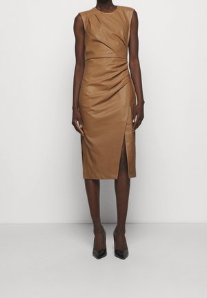 Robe midi en cuir marron sans manches avec fronces latérales et fente avant, portée avec des talons hauts noirs pointus sur un fond neutre.