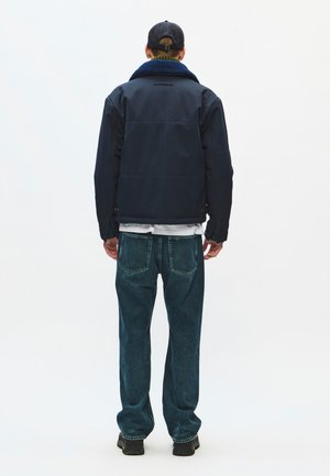 Mand iført en mørkeblå jakke med sherpa-krave, mørkeblå jeans, sorte sko og en sort baseballkasket, stående med ryggen mod på en ensfarvet baggrund.