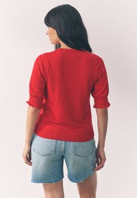 Woman with long dark hair facing backward, wearing a red short-sleeve sweater with ruffled cuffs and light blue denim shorts.