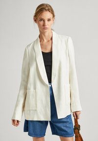 Blazer bianco in lino con apertura frontale, dotato di due tasche anteriori e maniche lunghe, abbinato a pantaloni corti in denim blu.