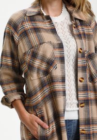 Rutig oversize skjortjacka i beige, brunt och blått med två bröstfickor, rundad krage och bruna knappar. Mjuk texturerad tyg.