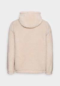 Veste en polaire beige avec capuche, texture douce et coupe décontractée. Épaules à côtes et ourlet sans fermetures visibles.