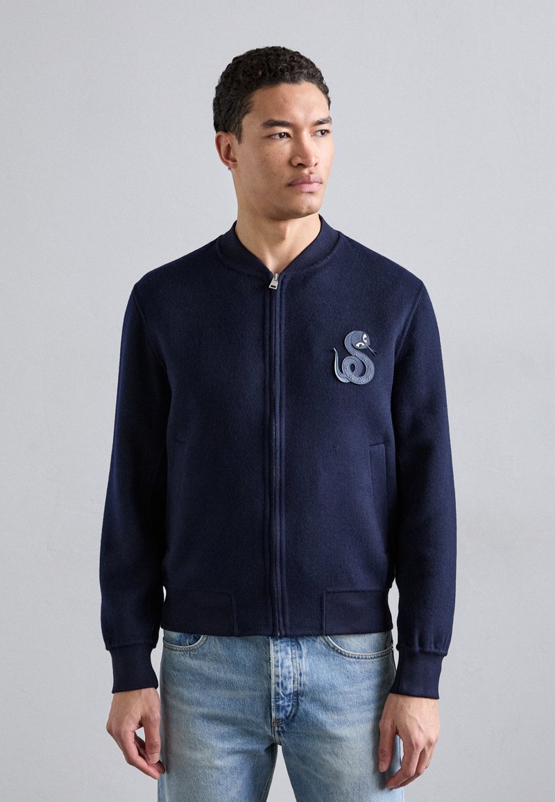 Jūrininkų mėlyna zip-up kardiganas iš minkštos, tekstūruotos medžiagos, su apvaliu apykakle, dviem priekinėmis kišenėmis ir gyvatės emblemą ant krūtinės.