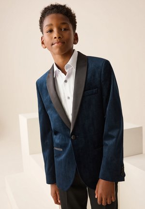 Jongen draagt een donkerblauwe fluwelen blazer met zwarte revers en een wit overhemd, staand tegen een effen beige achtergrond.