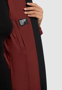 Mână ținând un sacou negru deschis, dezvăluind căptușeala roșie închisă, cu eticheta alb-negru "Khujo original for a few" în interior.