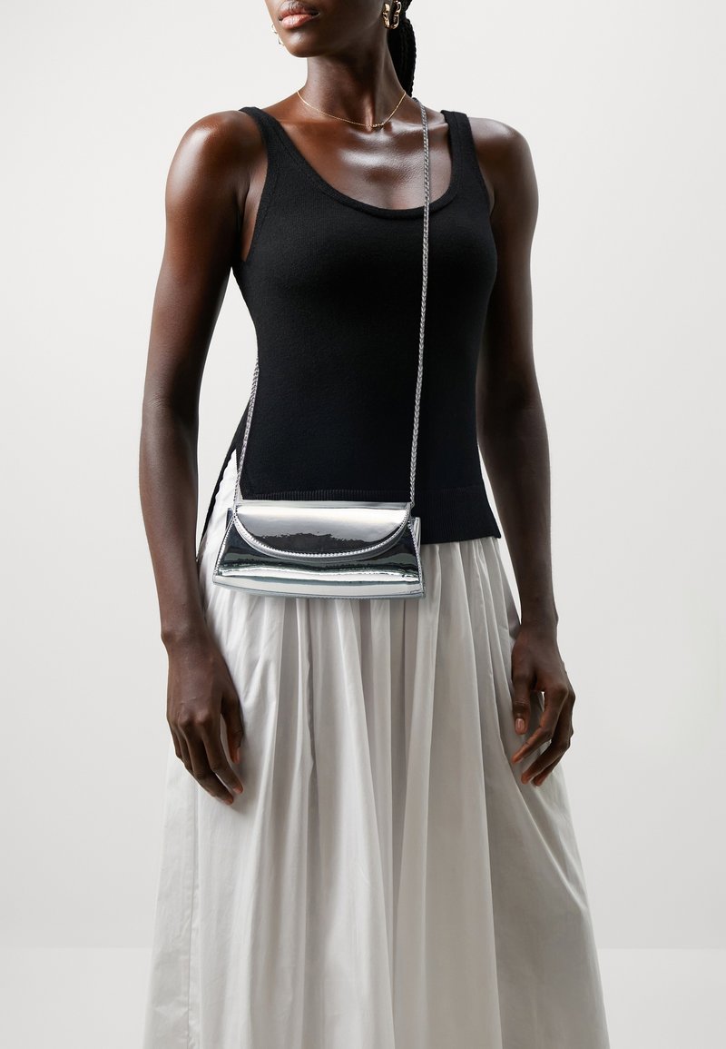 Femme portant un haut noir sans manches et une jupe blanche, portant un petit sac bandoulière argenté brillant avec une bandoulière en chaîne.