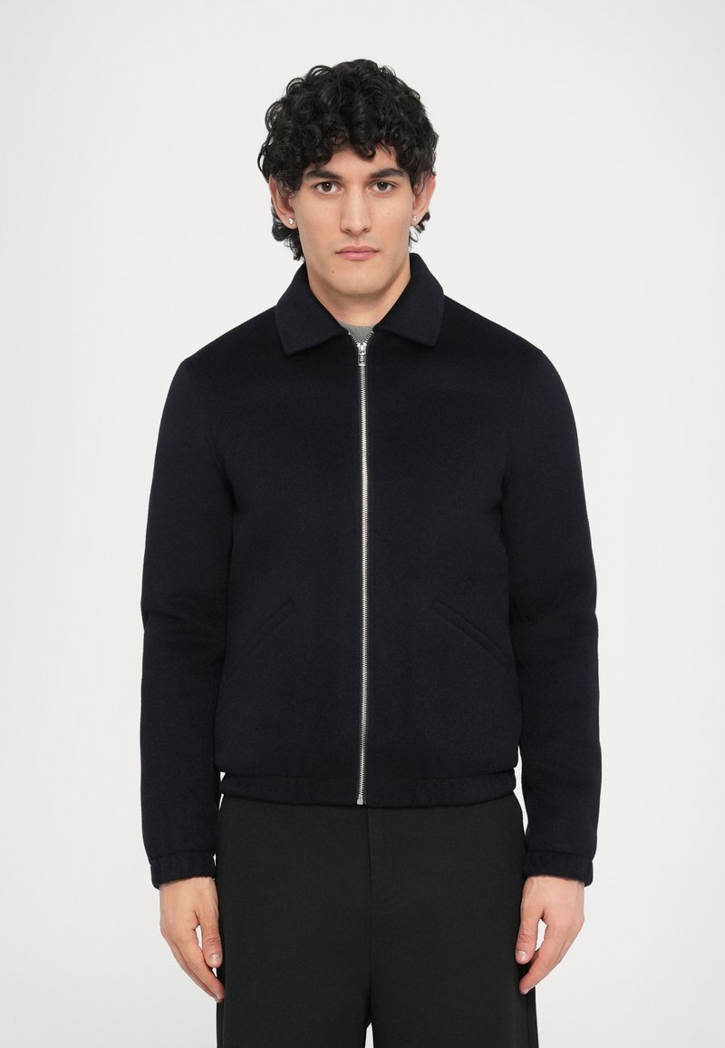 Tengerészkék gyapjúdzseki cipzáras elülső résszel, lehajtott gallérral és két oldalzsebbel. Bordázott mandzsetták és alsó szegély, sima textúrával.