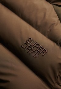 Brunt polstret stof med broderet "Super Dry®" logo i mørkebrunt tråd på overfladen.
