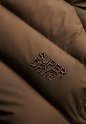 Brązowa, pikowana tkanina z haftowanym logo "Super Dry®" w ciemnobrązowej nici na powierzchni.