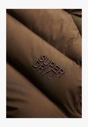 Brunt polstret stof med broderet "Super Dry®" logo i mørkebrunt tråd på overfladen.