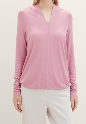 Femme portant un haut rose clair à manches longues et col en V, ainsi qu'un pantalon blanc, debout devant un fond clair uni.