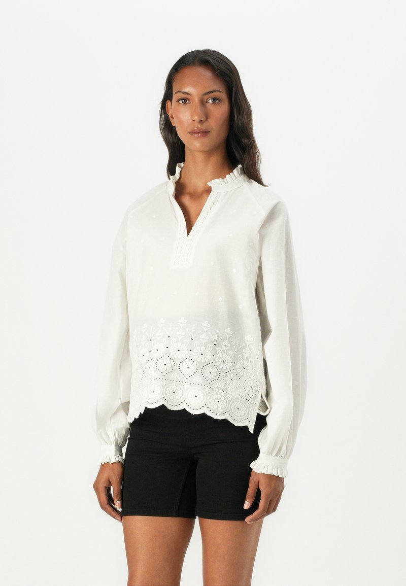 Hvit brodert bluse med bølget kant, med detaljerte blomstermønstre, v-hals med rynket krage og elastiske ermer.