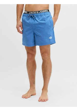 Uomo che indossa pantaloncini da bagno blu con vita elastica e coulisse, mani nelle tasche, in piedi scalzo su uno sfondo semplice.