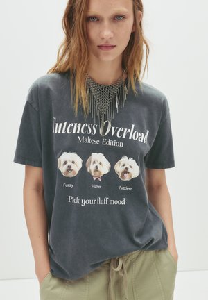 Grå skjorte med grafisk tryk, der viser tre Malteserhunde og teksten "Cuteness Overload Maltese Edition" nedenfor. Blødt stof, afslappet pasform.