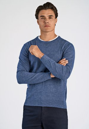 Herren blauer Strickpullover mit langen Ärmeln, rundem Ausschnitt, neutraler Textur, geripptem Saum, über einem weißen Shirt mit lässiger Passform getragen.