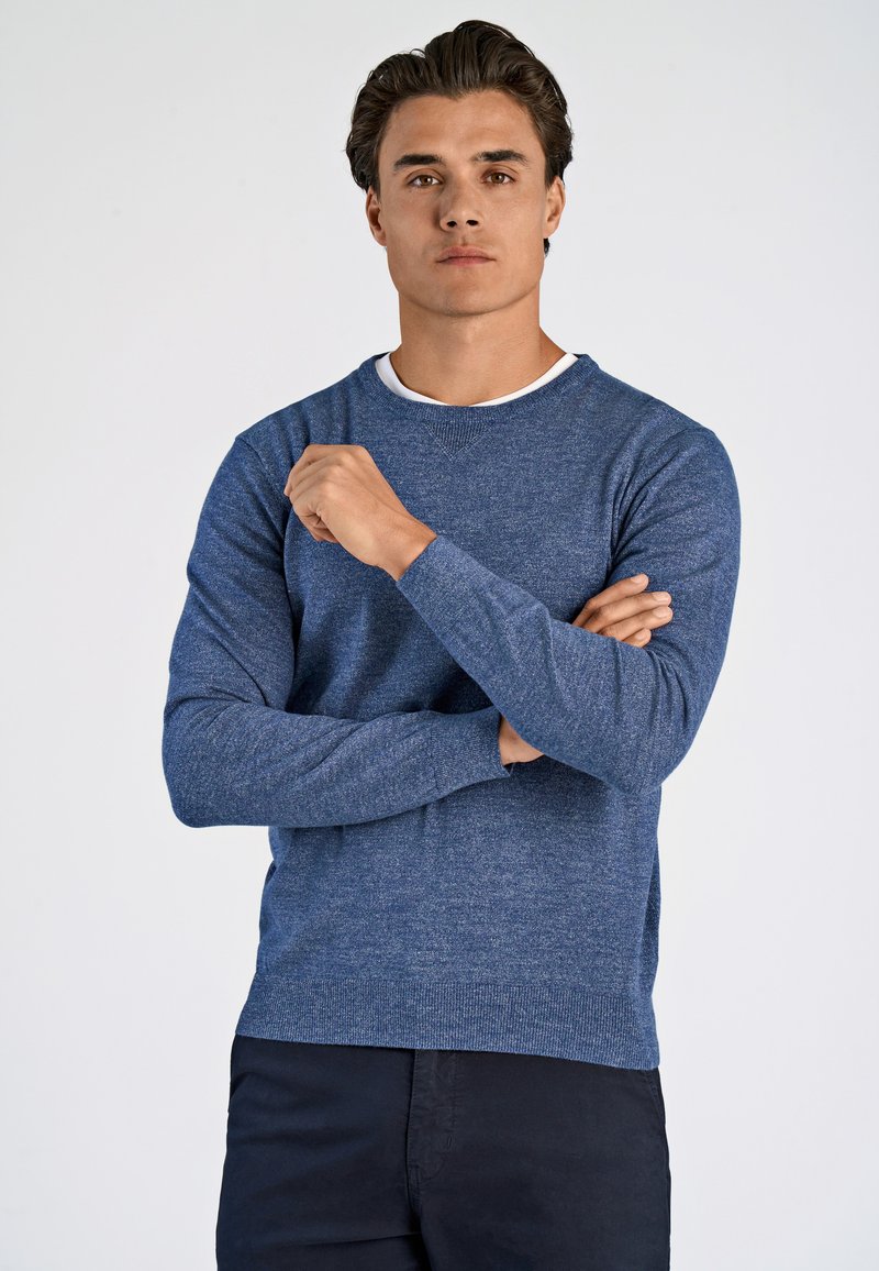 Herren blauer Strickpullover mit langen Ärmeln, rundem Ausschnitt, neutraler Textur, geripptem Saum, über einem weißen Shirt mit lässiger Passform getragen.