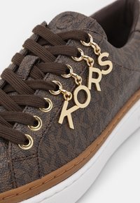 Sneaker marrone con texture e lacci marroni scuri infilati in occhielli dorati, con charm in oro a forma di lettere che spellano "KORS" sul lato.