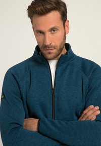 Blaues Zip-Jackett mit hohem Kragen, strukturierter Oberfläche und kontrastierenden schwarzen Bündchen. Verfügt über ein minimalistisches Logo auf dem linken Ärmel.