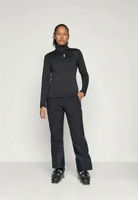 Μαύρη μπλούζα με μακριά μανίκια και ψηλό λαιμό, συνδυασμένη με μαύρα παντελόνια σκι. Και τα δύο διαθέτουν εφαρμοστή γραμμή και λεία ύφανση. Οι μαύρες μπότες σκι ολοκληρώνουν το σύνολο.