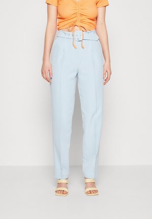 Femme portant un pantalon taille haute bleu clair avec ceinture, un top orange froncé à manches courtes et des sandales ouvertes jaunes, debout sur fond blanc.