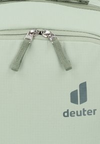 Grüner Stoffrucksack mit doppeltem silbernen Reißverschluss, strukturierter Oberfläche und grauem "deuter"-Logo. Die Reißverschlusszieher sind aus passendem grünem Stoff.