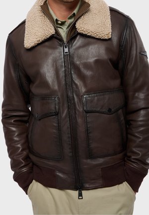 Mann trägt eine dunkelbraune Lederjacke mit beigem Lammfellkragen, vorderen Klapptaschen und gerippten Bündchen und Saum, über einem grünen Hemd.