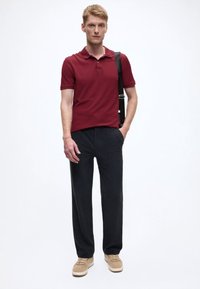 Burgundy polo shirt, dark fabric trousers, beige sneakers; model stands with one hand in pocket, minimalist background.