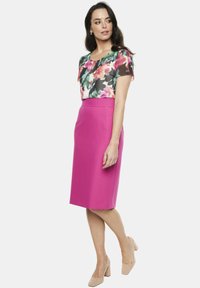 Bluzka w kwiatowy wzór w bieli, różu i zieleni, zestawiona z dopasowaną różową spódnicą ołówkową. Modelka ma na sobie cieliste buty na obcasie.