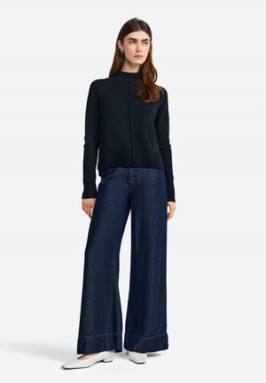 Zwarte gebreide trui met hoge hals, gecombineerd met wijde donkerdenim broeken en witte puntige flats. Soepele textuur, minimalistisch ontwerp.
