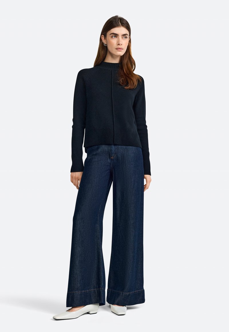 Black knit sweater with high neck, paired with wide-leg dark denim pants and white pointed-toe flats. Smooth texture, minimal design.