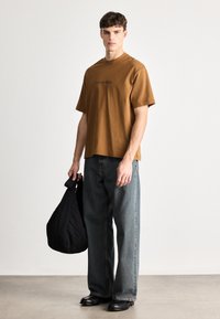 Καφέ βαμβακερό μπλουζάκι με λογότυπο Calvin Klein, συνδυασμένο με φαρδιά μπλε τζιν παντελόνια και μαύρα υποδήματα. Κρατάει μια μαύρη τσάντα ώμου.