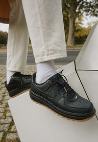 Baskets en cuir noir à finition texturée, détails en métal doré, lacets noirs et semelle en caoutchouc gomme, portées avec des chaussettes blanches et un pantalon clair.
