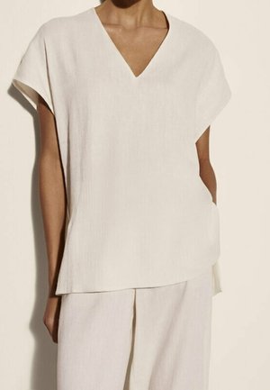 Femme portant un haut crème ample à manches courtes et encolure en V accompagné d'un pantalon large assorti, sur un fond uni.
