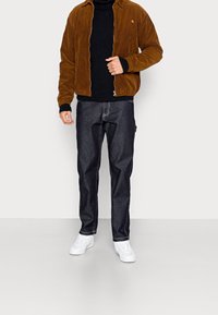 Brązowa kurtka z kordurowego materiału z zamkiem z przodu, czarny sweter dzianinowy pod spodem, ciemne dżinsy i białe trampki. Prosty, nowoczesny design.