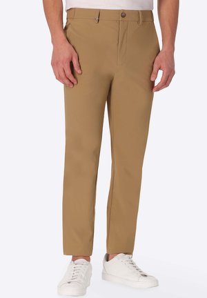 Uomo con pantaloni beige a gamba dritta e scarpe da ginnastica bianche in piedi davanti a uno sfondo semplice.
