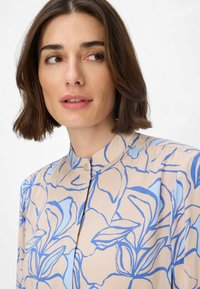 Blusa beige chiaro con disegni floreali blu a linea, caratterizzata da un colletto alto e una chiusura con bottoni. Tessuto morbido con un design fluido.