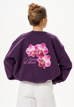 Vrouw met haar in een knot draagt een paarse sweatshirt met roze orchideeënbloemen en de tekst "Bloom in silence" op de rug, gecombineerd met een witte broek.