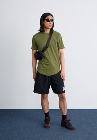 Un jeune homme se tient contre un mur neutre, portant des lunettes de soleil, un t-shirt vert, un short noir et des baskets, avec un petit sac noir en bandoulière.