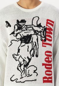 Witte gebreide trui met een zwart rodeo grafiek en de tekst "Rodeo Town" in het rood. De textuur is lichtgewicht en los geweven.