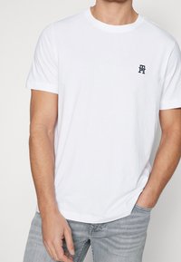 T-shirt in cotone bianco con scollo rotondo e maniche corte, con un piccolo logo blu navy sul lato sinistro del petto. Vestibilità casual.