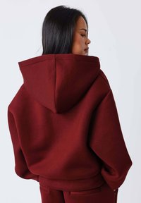 Sudadera con capucha color burdeos hecha de tela suave, con un corte relajado, puños acanalados y detalles de costura en la espalda. Ideal para uso casual.