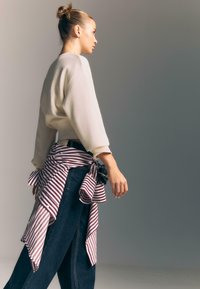 Žena s vlasmi v bunke, oblečená v bielom svetri a džínsach, s pruhovanou košeľou zviazanou okolo pásu, kráčajúca proti šedému pozadiu.