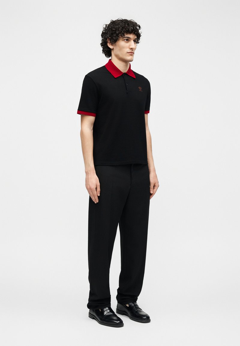 Man in black pants and black polo shirt with red collar and sleeve edges, standing in neutral pose against plain background.