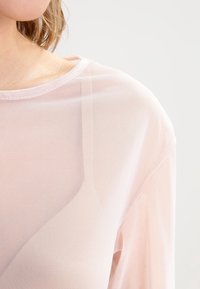 Světle růžový průsvitný top vyrobený z jemné síťoviny, s kulatým výstřihem a volným střihem, který ukazuje jemnou strukturu a hladký povrch.