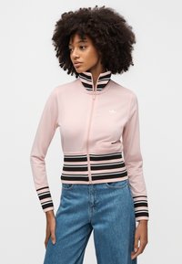 Ροζ μπουφάν με φερμουάρ και ψηλό γιακά, με οριζόντιες μαύρες και άσπρες ρίγες στη μέση και στα μανίκια. Λευκό λογότυπο στο στήθος.